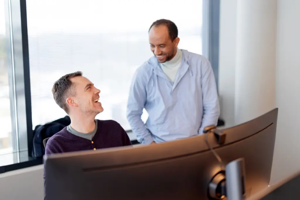 To menn som snakker sammen foran en stor dataskjerm. Den ene sitter og den andre står