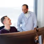 To menn som snakker sammen foran en stor dataskjerm. Den ene sitter og den andre står