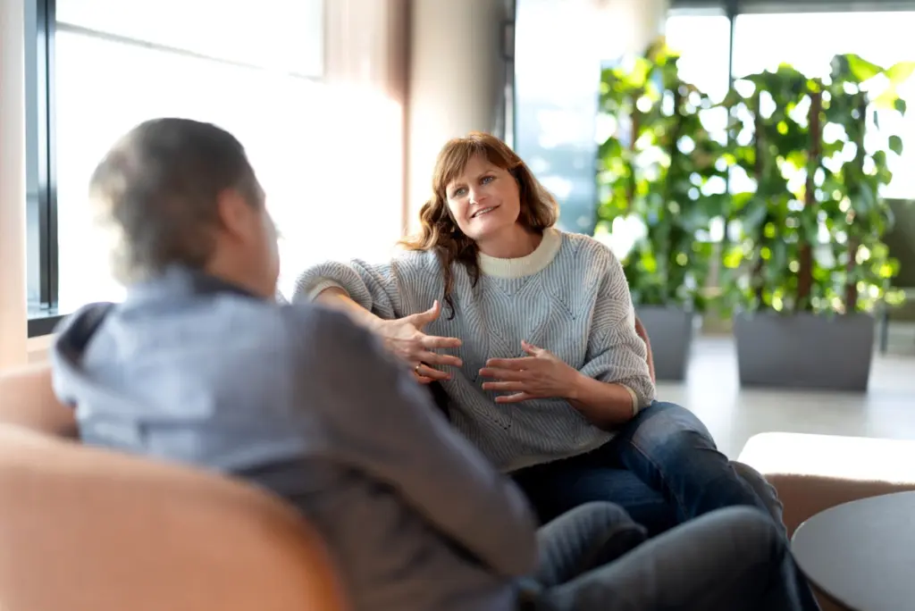 To personer, en mann med ryggen til og en dame som ser mot kamere, sitter i en sofa og snakker sammen.