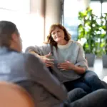 To personer, en mann med ryggen til og en dame som ser mot kamere, sitter i en sofa og snakker sammen.