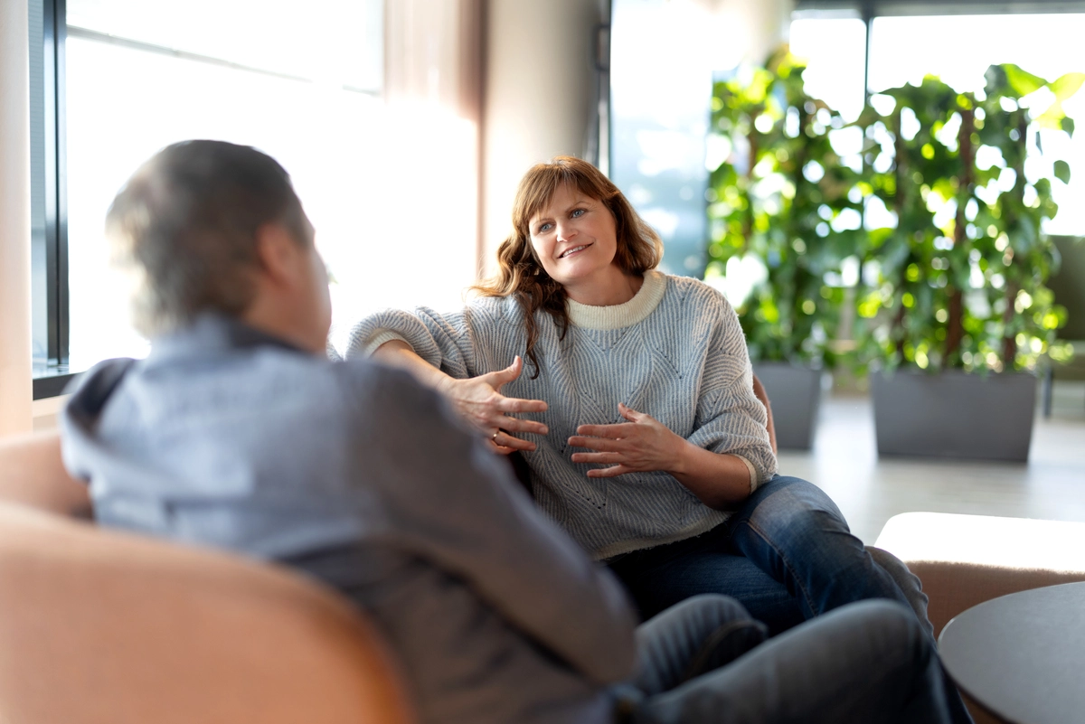 To personer, en mann med ryggen til og en dame som ser mot kamere, sitter i en sofa og snakker sammen.